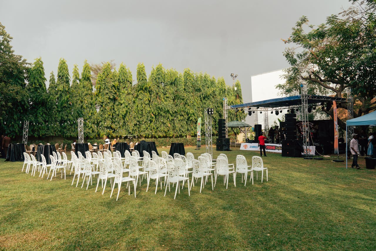 An outdoor event venue with white chairs, stage setup, sound equipment, and greenery surrounding the space.